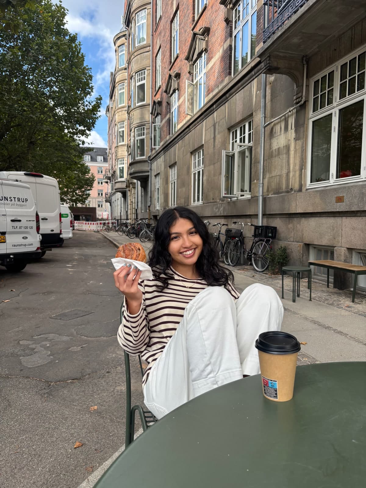 Sadia at a sidewalk cafe table, smiling with a pastry in one hand and a coffee beside her